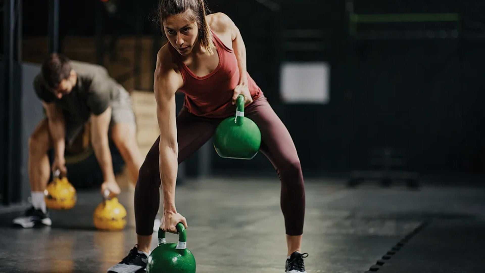 Woman lifting green kettlebells in a gym setting with another person in the background.