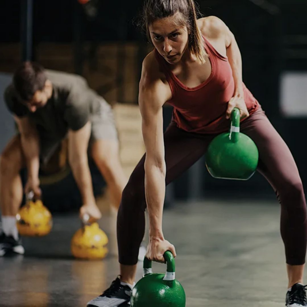 Woman lifting green kettlebells with another person in the background at a gym.