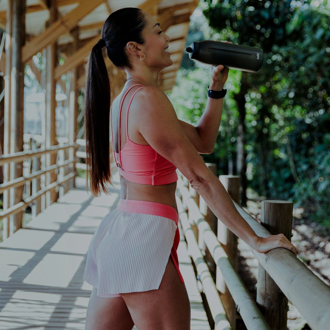 Woman in athletic wear drinking from a water bottle on a wooden deck with greenery.